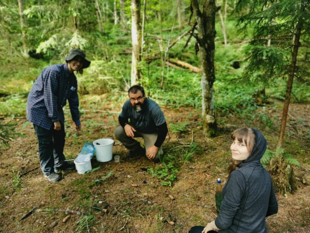 Kolme ihmistä ovat metsässä ja katsovat iloisena kameraan. Kaksi ihmisistä on kyykistyneenä, yksi seisoo ämpäreiden vieressä.